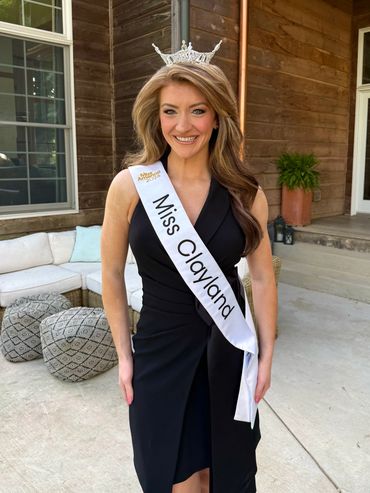 A smiling woman wearing a Miss Clayland sash and crown in a black dress.