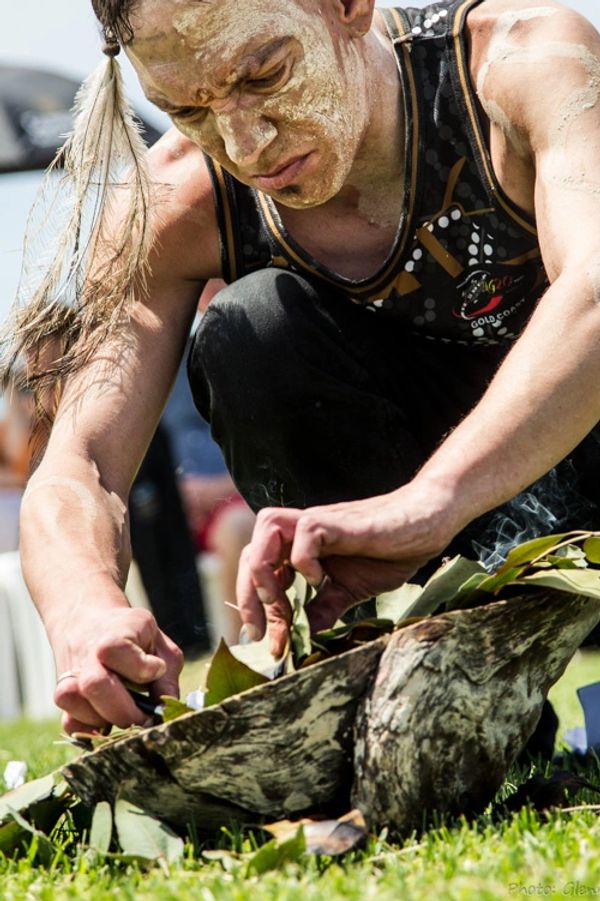 A man with mud on his face prepares a traditional fire using leaves and bark outdoors.