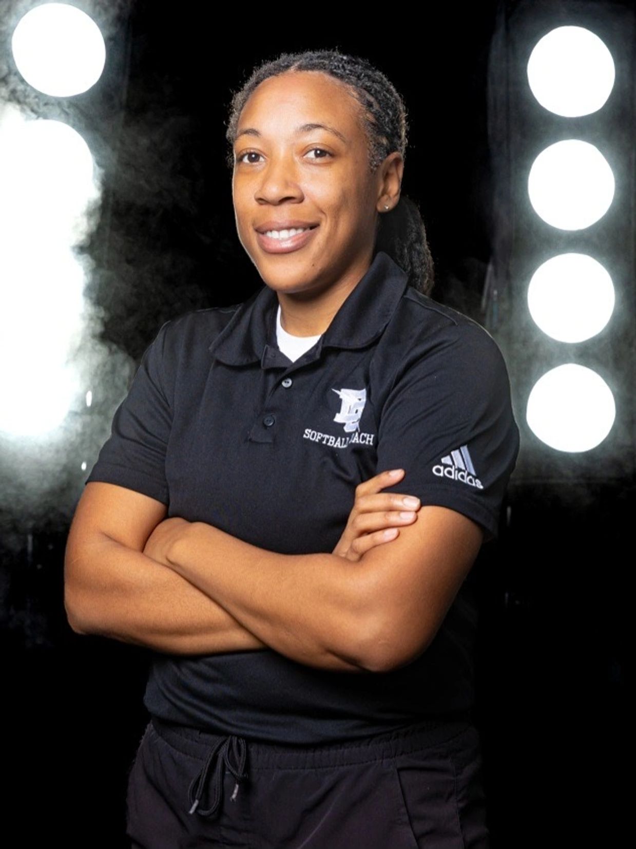 A confident woman in a black softball polo shirt stands with arms crossed, smiling against a backdrop of bright circular lights.