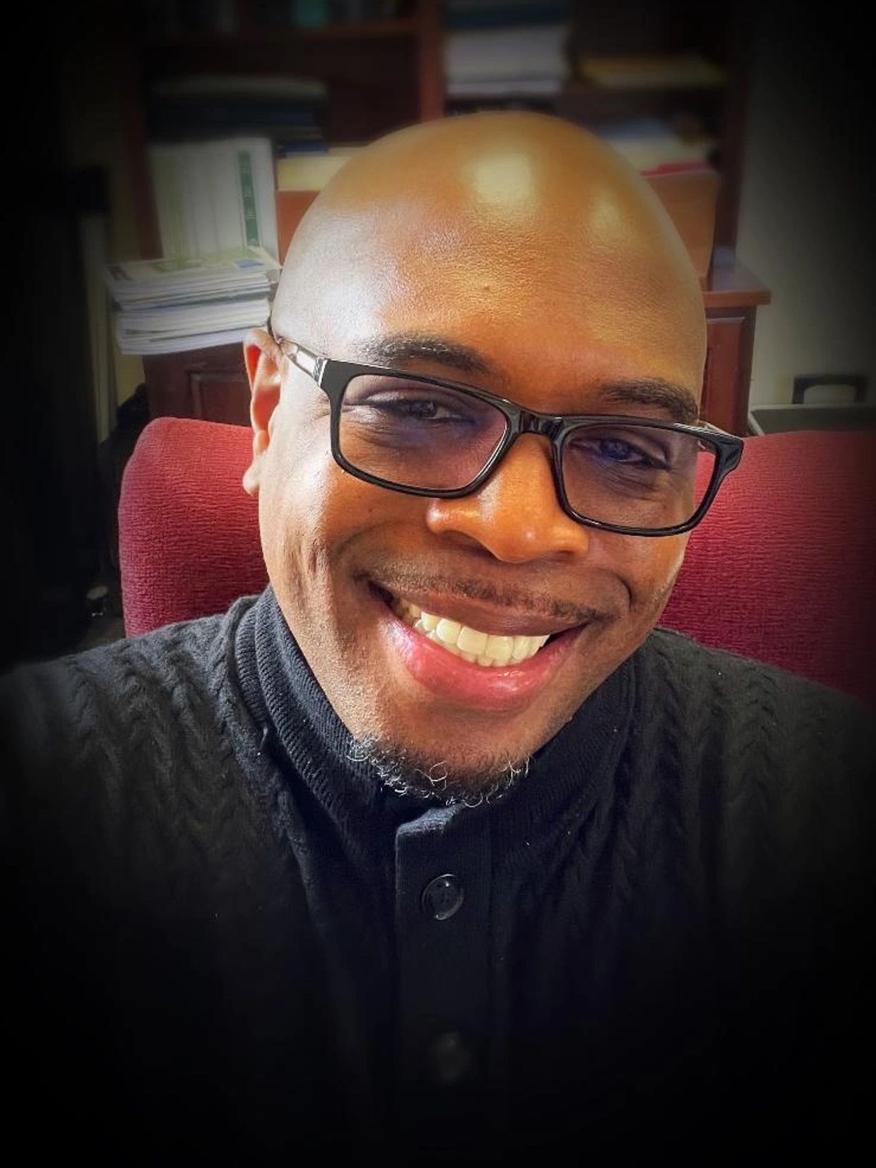 Smiling man wearing glasses and a black sweater, sitting in a red chair.