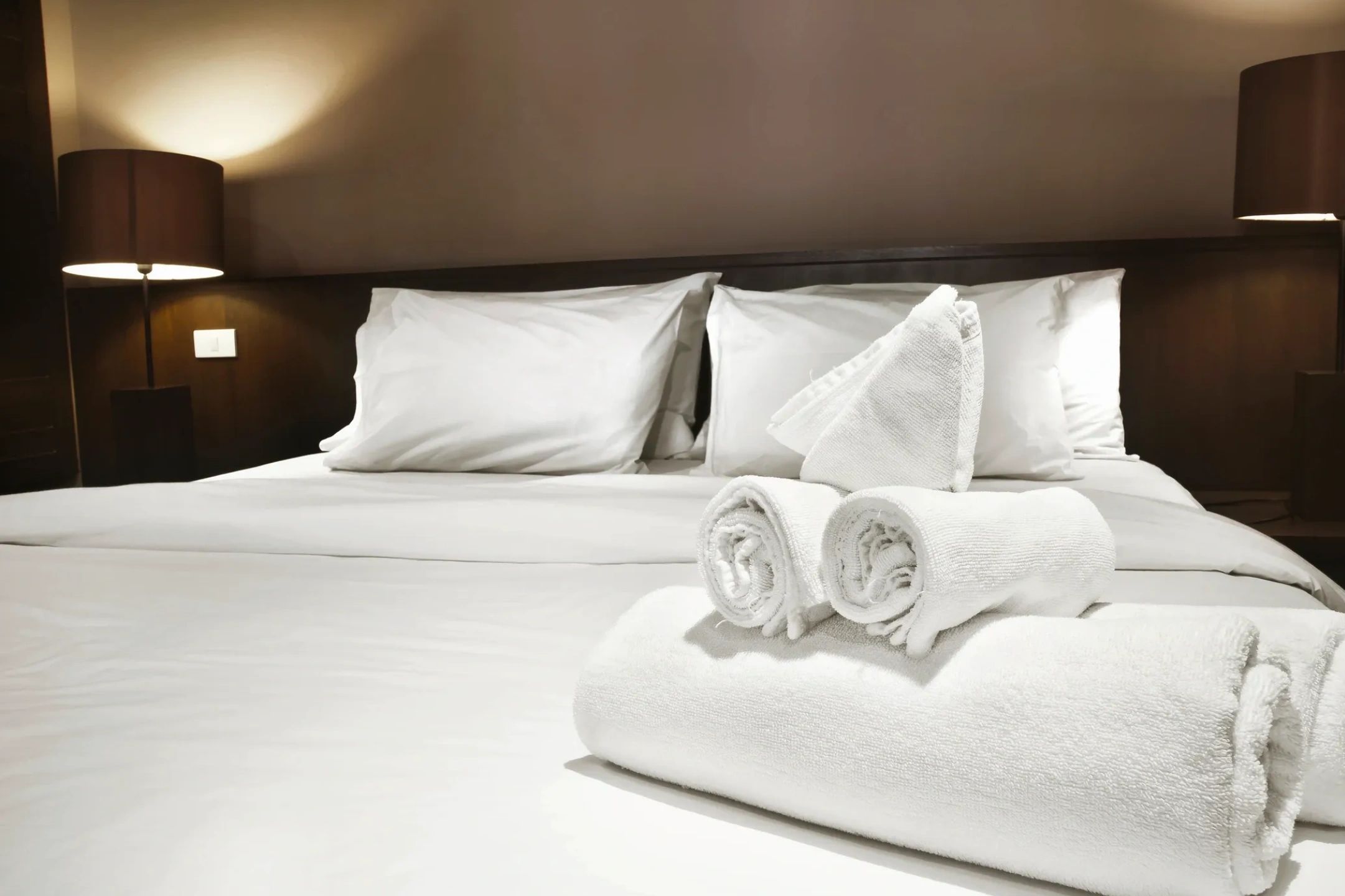 Neatly arranged white towels on a bed with white pillows and bedside lamps.