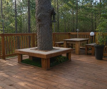 Wooden deck with built-in seating around a tree and a picnic table surrounded by forest.