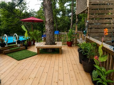 A wooden deck with plants, a pool, and a tree bench under a red umbrella.