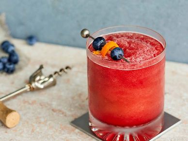 Red cocktail garnished with blueberries and orange peel on a coaster.