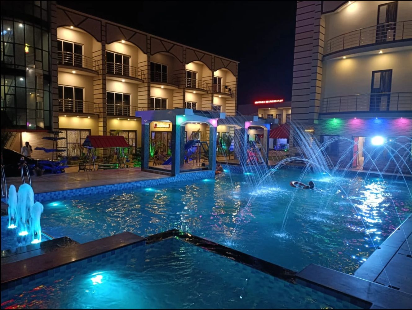 Night view of a lit swimming pool with fountains and hotel buildings.