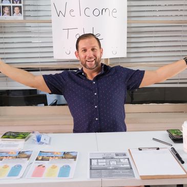 Smiling man welcomes guests at a table with informational brochures and a sign.