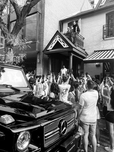 Crowd gathered around a man on a balcony, many taking photos, with a black Mercedes parked nearby.