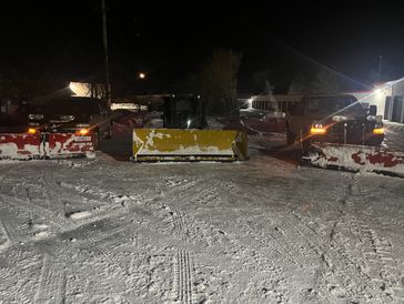 Three snowplows parked side by side in a snowy area at night.