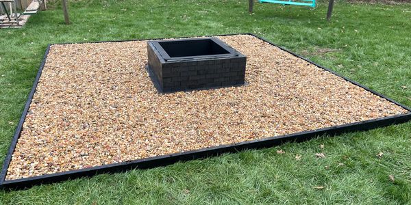 Square fire pit on a bed of small rocks in a grassy yard.
