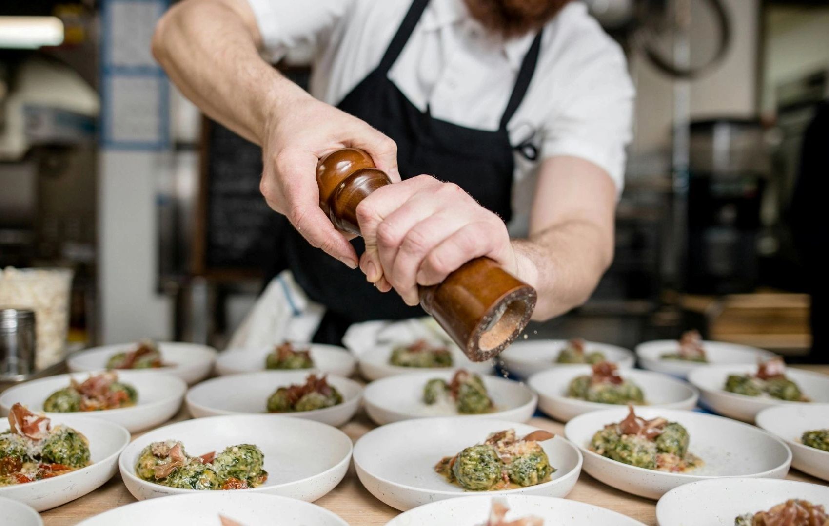 Chef grinds pepper over plates of gourmet spinach dumplings in a busy kitchen.