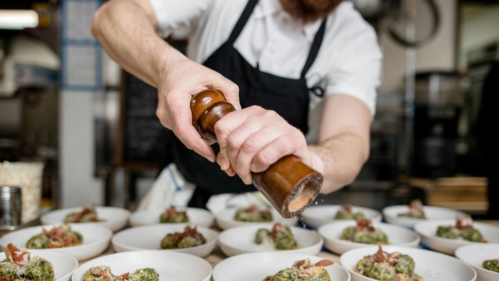 Chef grinds pepper over plates of gourmet spinach dumplings in a busy kitchen.