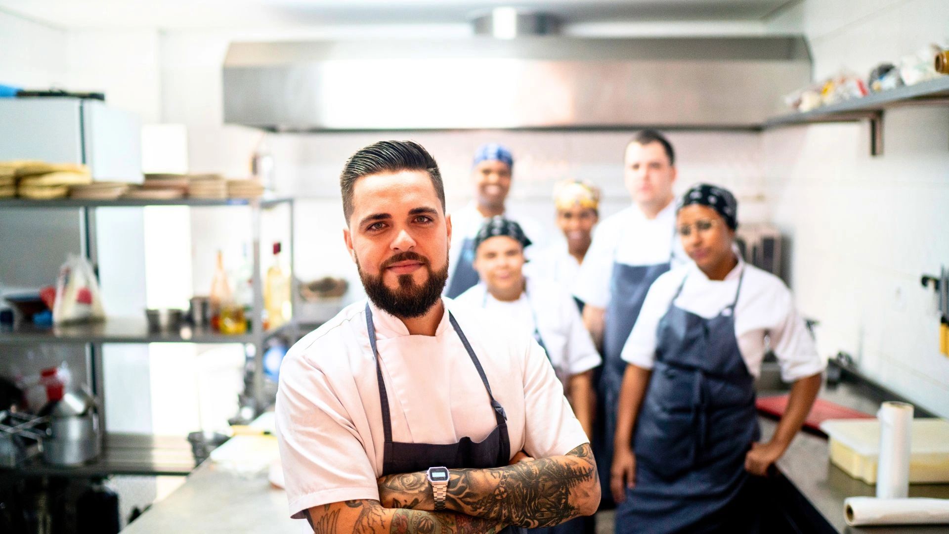 A confident chef with tattoos stands in front of a diverse kitchen team.