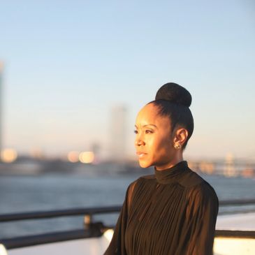 A woman in a black dress looks thoughtfully over a waterfront at sunset.