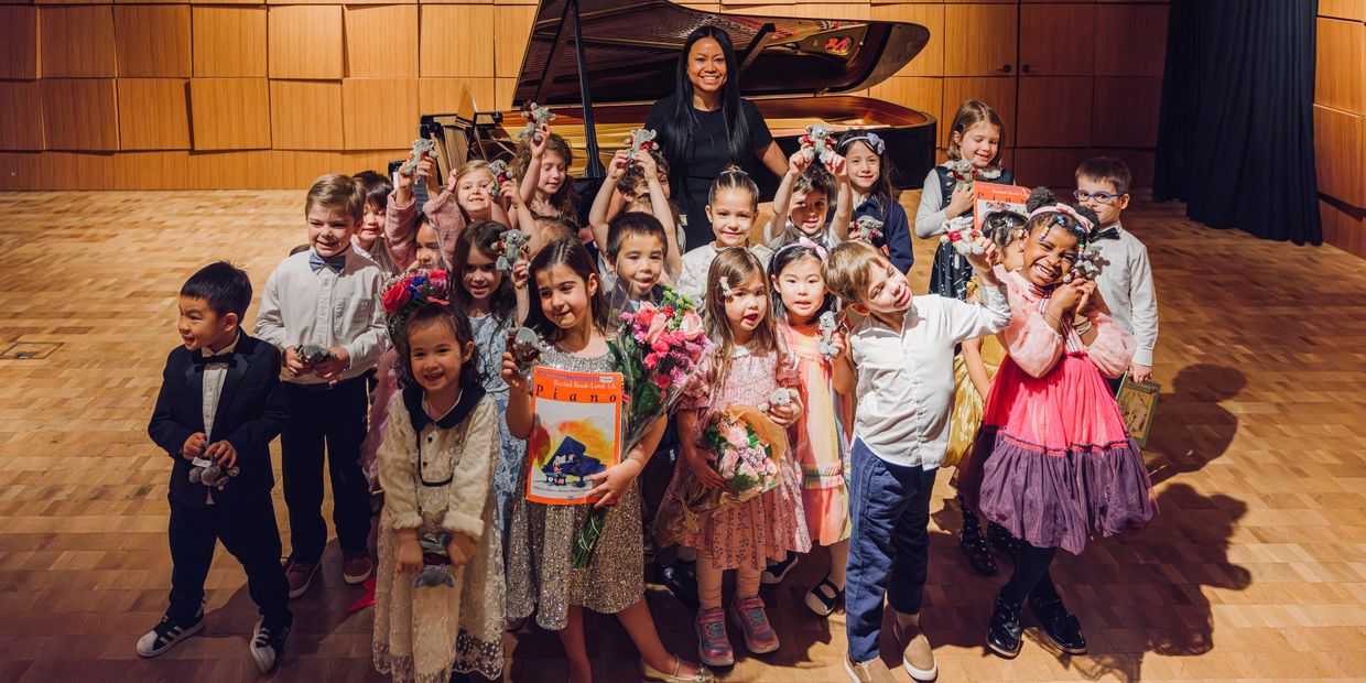 A group of children with their teacher posing on stage after a piano recital.