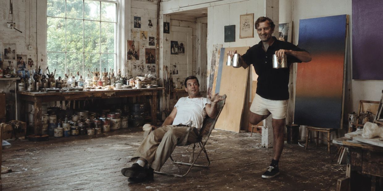 Two men in a cluttered art studio, one sitting and the other standing with paint cans.