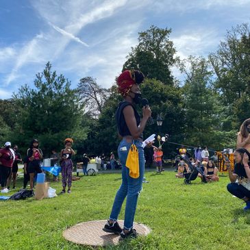 A woman speaks with a microphone at a vibrant outdoor community gathering.