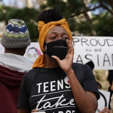 A woman wearing a mask speaks at a protest with signs in the background.