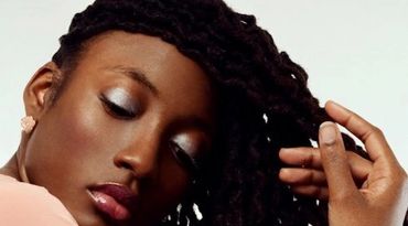 A woman with dark skin and twisted hair, eyes closed, gently touching her hair.