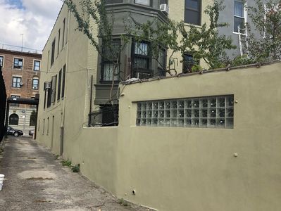 Alley beside beige building with glass block window and greenery.