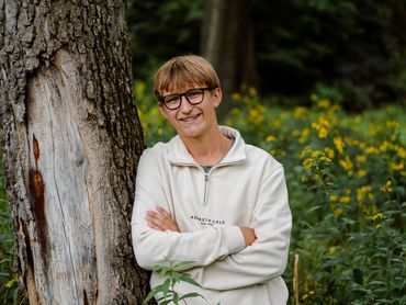 Young man with glasses leaning against a tree in a green, flowery outdoor setting.