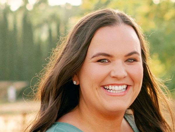 A woman with long brown hair smiling outdoors in natural light.
