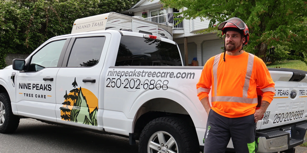 A tree care worker in safety gear stands next to a branded company truck.