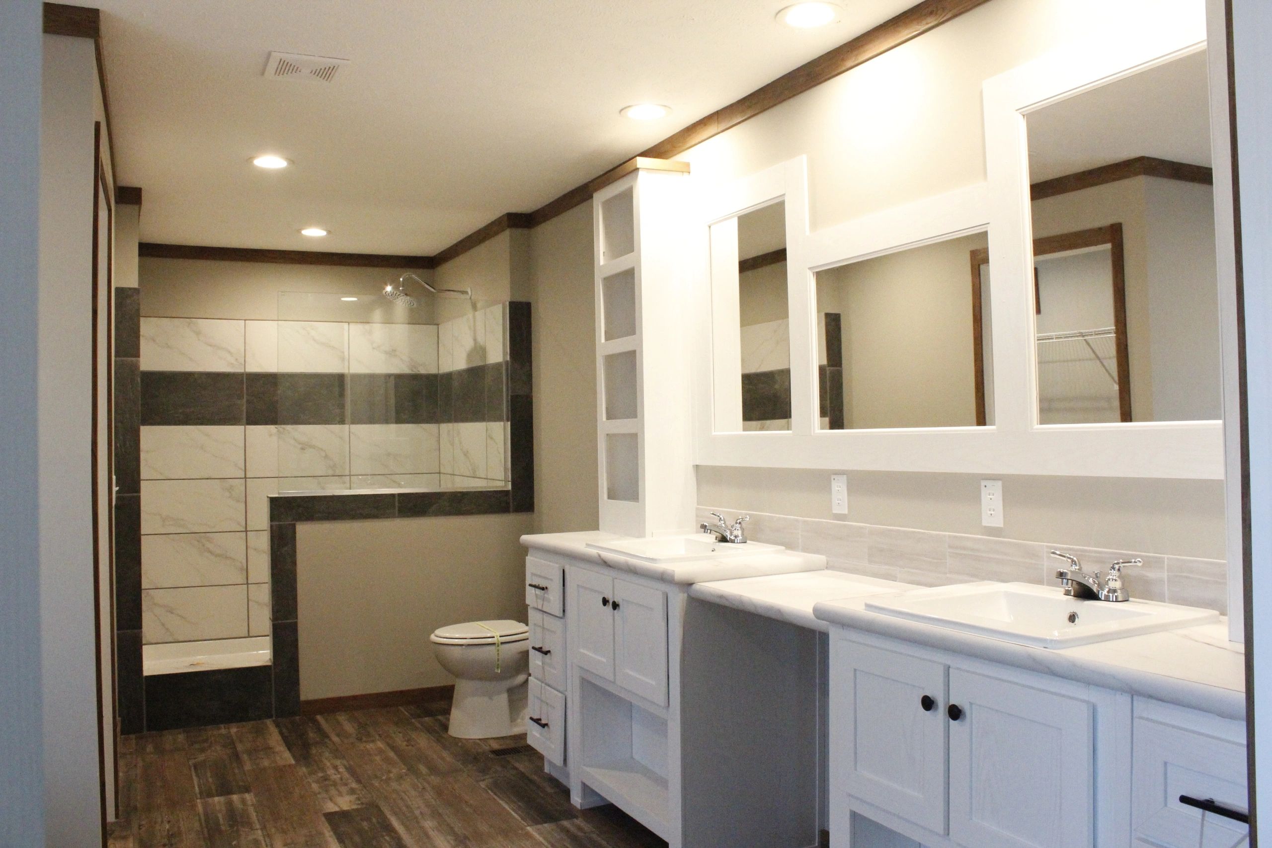 Spacious bathroom with dual sinks, large mirrors, and a walk-in shower on the left. Warm wood flooring and neutral tones create a modern, serene feel.