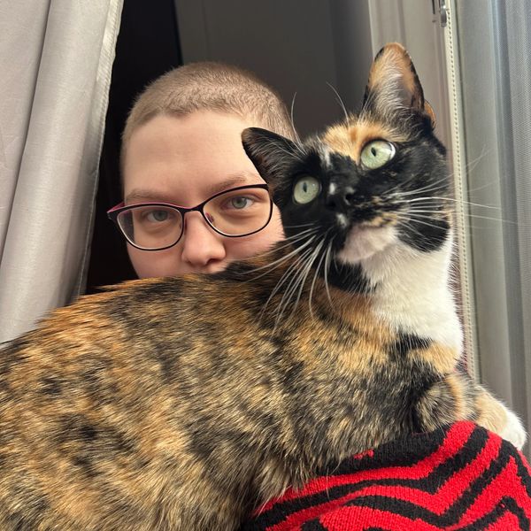 Person with glasses and short hair behind a calico cat near a window.