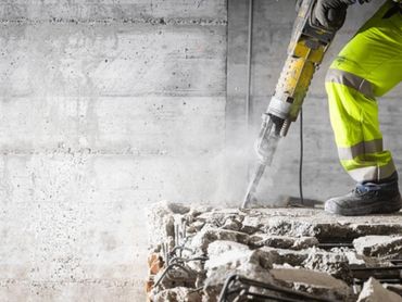 Worker using jackhammer to break concrete with dust flying around.