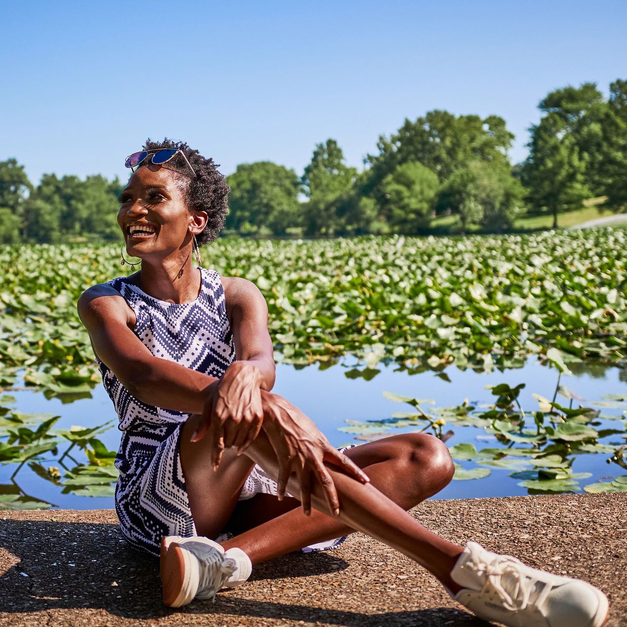 Black female smiling & sitting on concrete. A pond of water with a sea of lily pads is behind her.