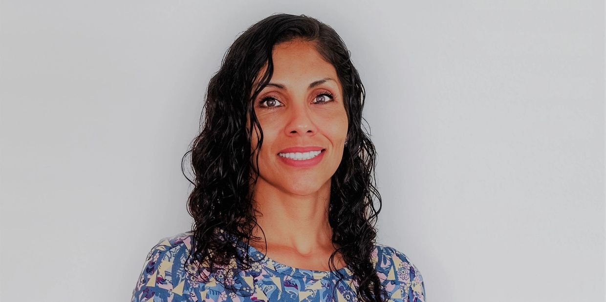 Woman in a patterned blue dress sitting and smiling against a plain background.