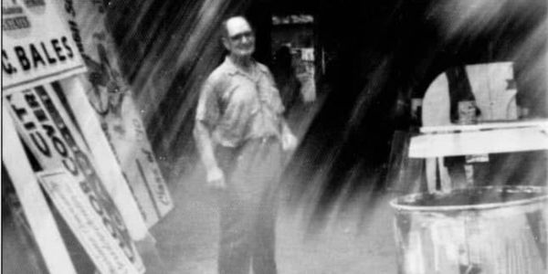 Black and white photo of a man standing near storefront signs.