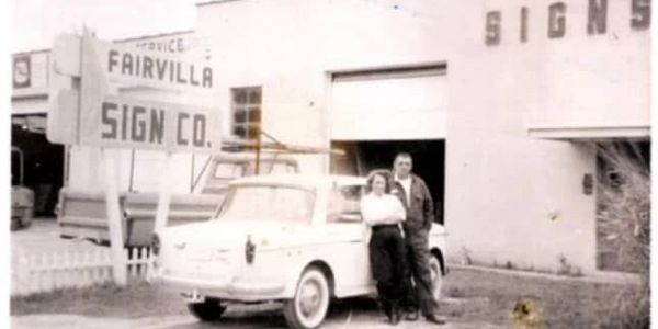 A vintage photo of a couple standing by a classic car outside Fairvilla Sign Co.
