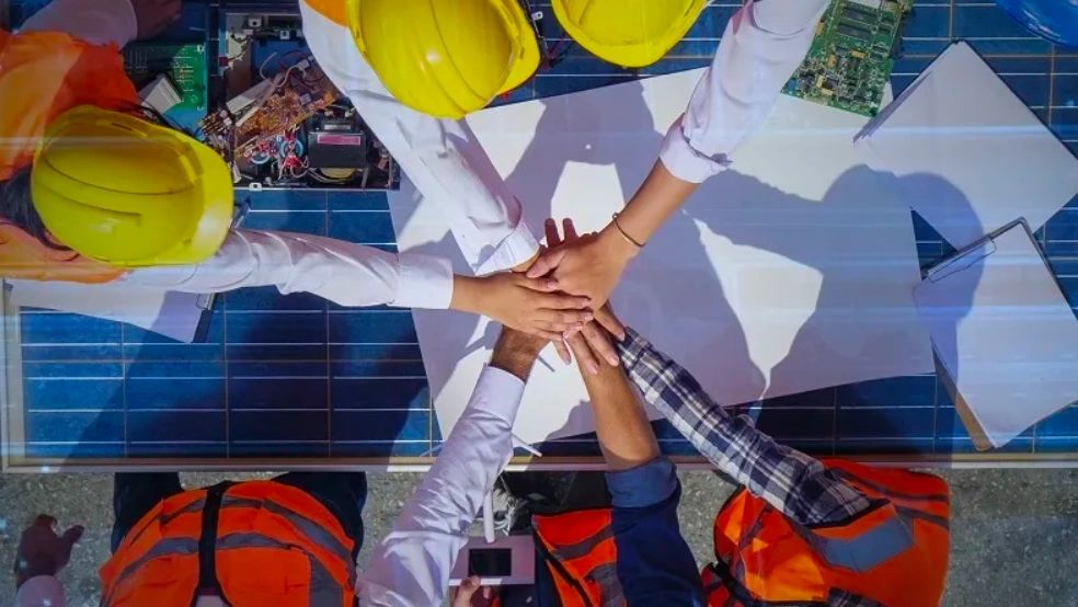 Construction workers with helmets join hands in teamwork over blueprints.