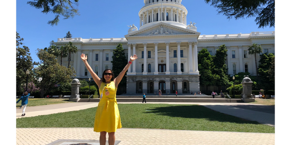 Dr Cio wearing a yellow dress, raising her arms and smiling in front of a government building.