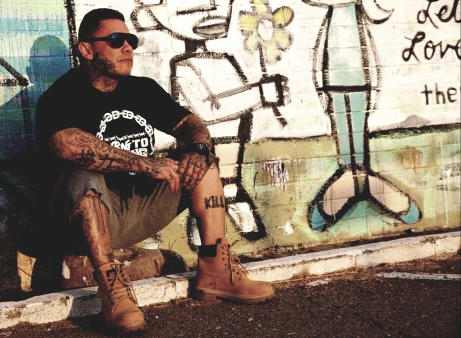 Tattooed man sitting by a graffiti wall.