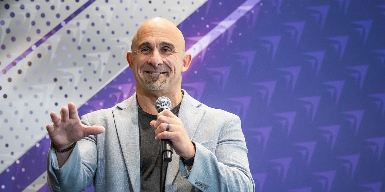 Man in a light gray blazer speaking with a microphone against a purple patterned background.