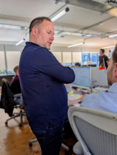 A man in a navy shirt stands and talks to a seated coworker in a busy office.