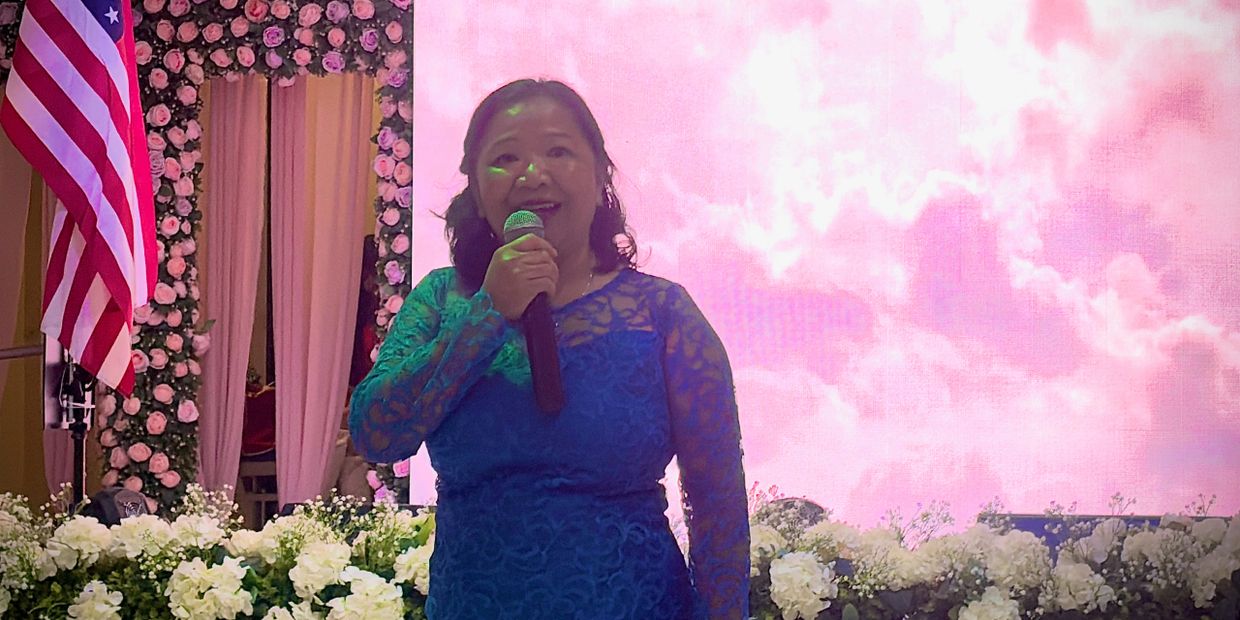 Woman in blue lace dress speaking into a microphone at a decorated event.