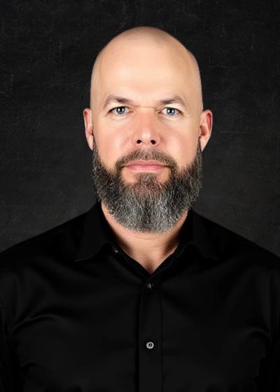 Portrait of a bald man with a gray beard wearing a black shirt against a dark background.