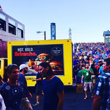 Crowd outside a venue with a bright Sriracha food truck in the foreground.
