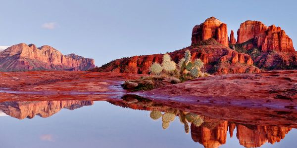 Red rock formations and cactus reflected in calm water under a clear sky.