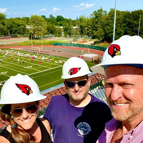 Three people wearing white hard hats with cardinal logos at a football field.