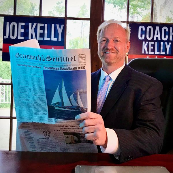Man in suit holding a newspaper with Joe Kelly campaign signs behind him.
