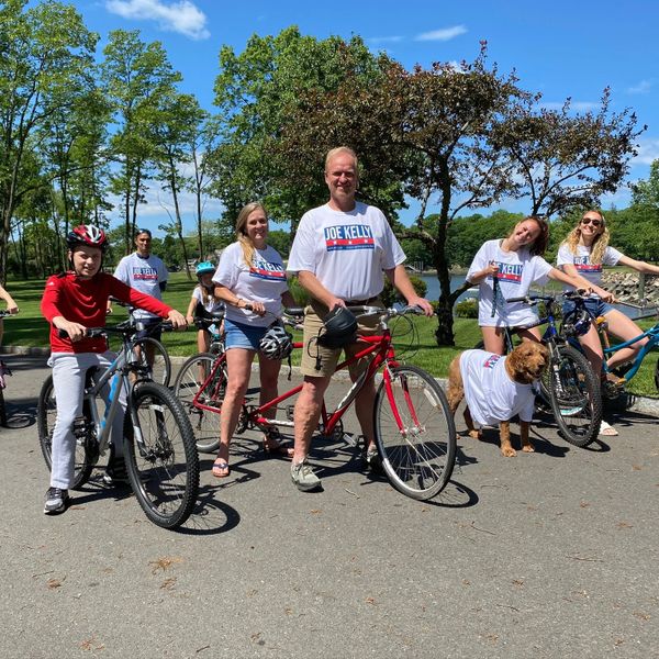Group of people and a dog wearing Joe Kelly shirts with bikes outdoors.