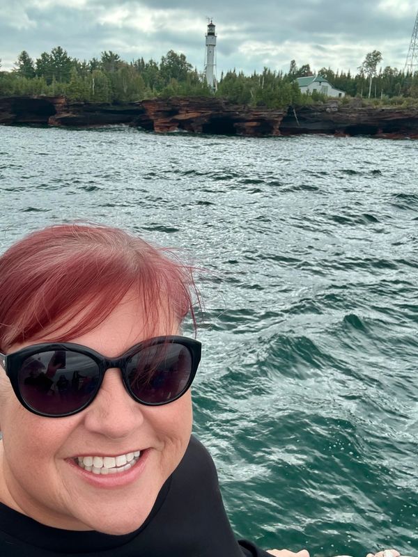 Woman smiling wearing sunglasses near water with lighthouse in background.