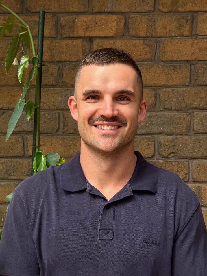 Smiling man with a mustache wearing a navy polo shirt against a brick wall.