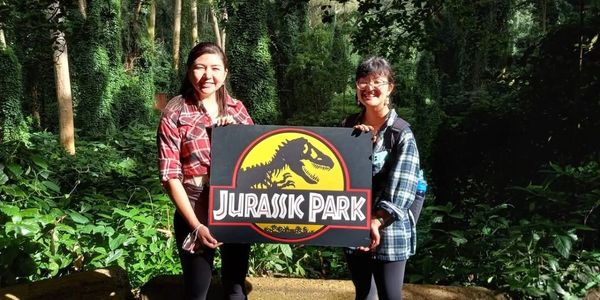 Two women holding a Jurassic Park sign in a forest.