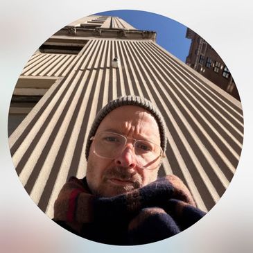 Man wearing glasses and a beanie poses in front of a tall building with vertical lines.