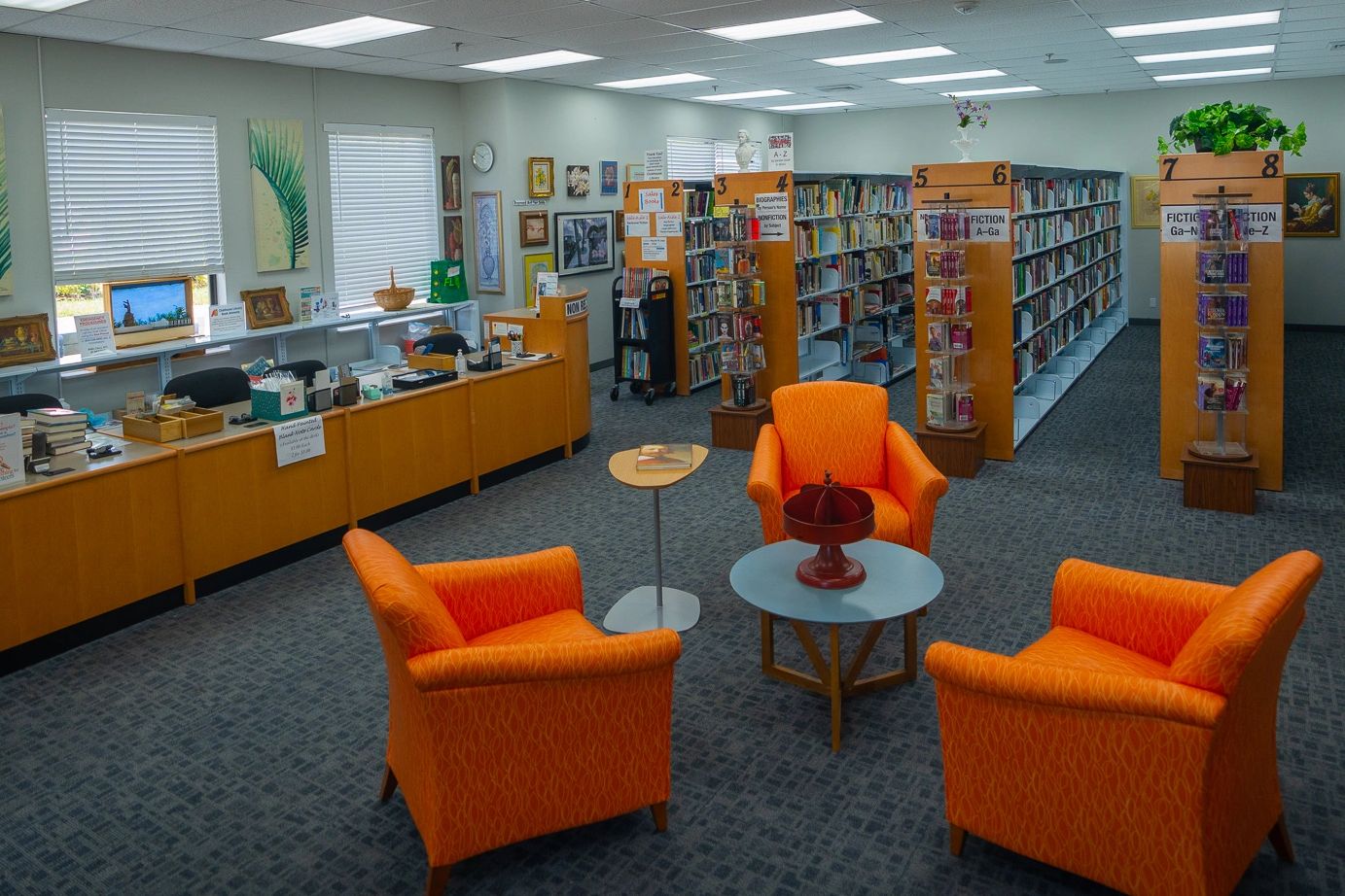 Bright orange chairs arranged around tables in a quiet library section with bookshelves and a checkout desk.